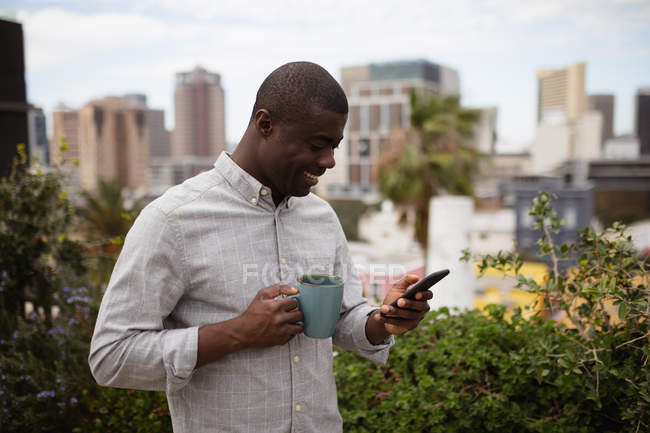 Вид збоку крупним планом молодого афроамериканського людини, що стоїть на вулиці на балконі в місті тримає чашку кави і дивлячись на його смартфон посміхаючись — стокове фото