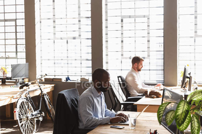 Seitenansicht eines jungen afrikanisch-amerikanischen Mannes und eines jungen kaukasischen Mannes, die in einem kreativen Büro am Schreibtisch sitzen und Computer benutzen — Stockfoto