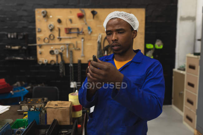 Vista frontal de perto de um jovem trabalhador de fábrica afro-americano que inspeciona equipamentos na loja de máquinas em uma fábrica de processamento, com equipamentos e ferramentas em segundo plano — Fotografia de Stock