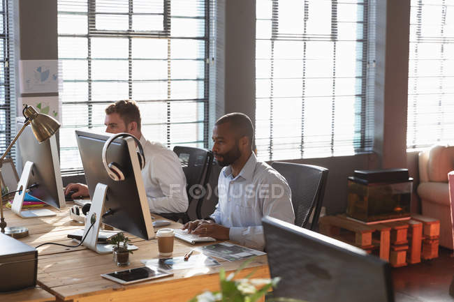 Vue latérale d'un jeune homme afro-américain et d'un jeune homme caucasien assis à un bureau à l'aide d'ordinateurs dans un bureau créatif — Photo de stock