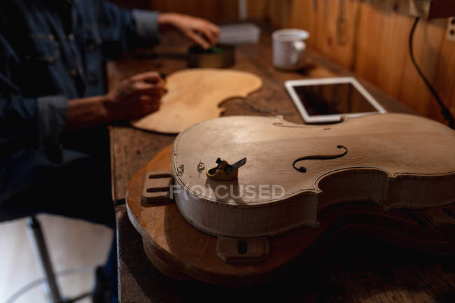 Vista lateral del luthier femenino trabajando en el cuerpo de un violín con una tableta en un banco de trabajo - foto de stock