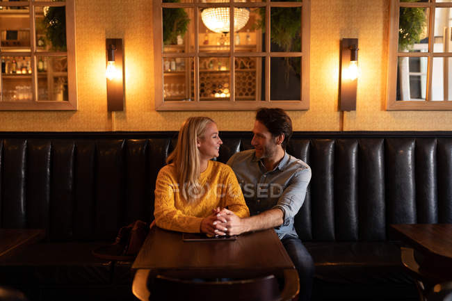 Vista frontal de una feliz pareja caucásica joven relajándose juntos en vacaciones en un bar, abrazándose y tomándose de la mano - foto de stock