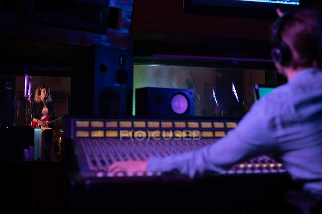 Visão traseira de um jovem engenheiro de som caucasiano sentado e trabalhando em uma mesa de mistura, com uma jovem músico caucasiana tocando guitarra elétrica em uma sala de gravação vista pela janela em um estúdio de gravação — Fotografia de Stock