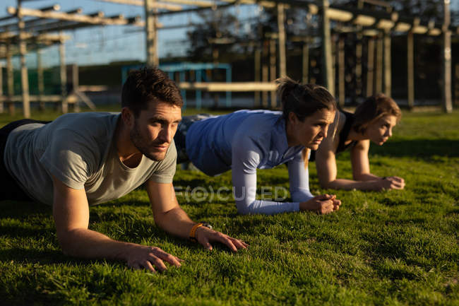 Vista lateral de duas jovens mulheres caucasianas e um jovem caucasiano fazendo o exercício de prancha em um ginásio ao ar livre durante uma sessão de treinamento de bootcamp — Fotografia de Stock