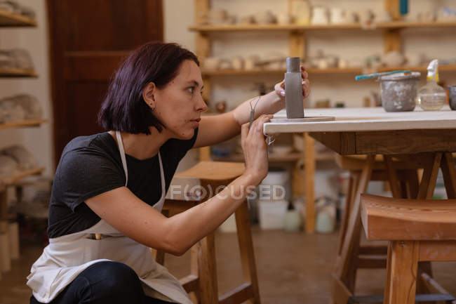 Side view of a young Caucasian female potter kneeling down and holding a clay flask on a table and checking it — Stock Photo