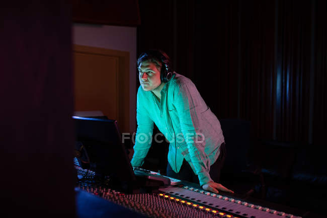 Vista lateral de cerca de un joven ingeniero de sonido caucásico de pie en un escritorio de mezcla en un estudio de grabación con auriculares - foto de stock