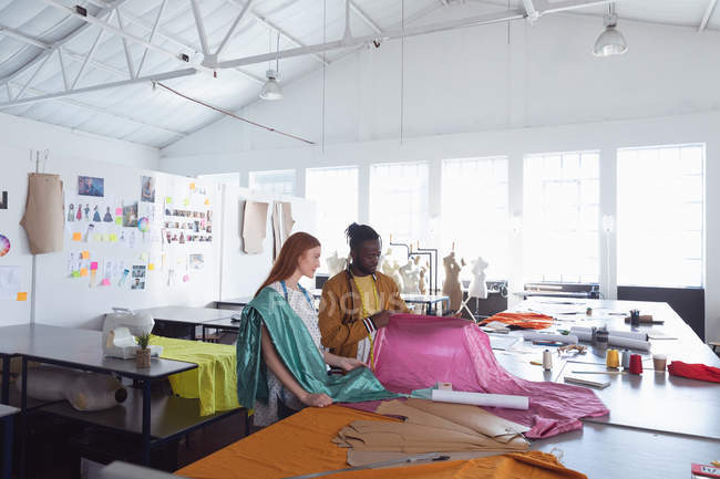 Vista lateral de uma jovem estudante de moda feminina caucasiana e uma jovem estudante de moda masculina afro-americana olhando para tecidos rosa e azul enquanto trabalhava em um projeto em um estúdio na faculdade de moda — Fotografia de Stock