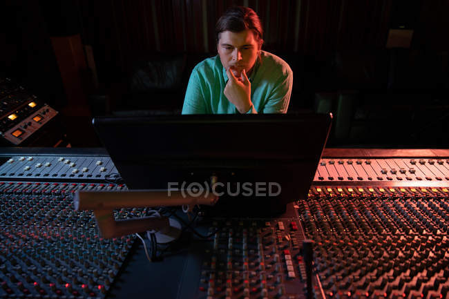 Front view of a young Caucasian male sound engineer sitting and working at a mixing desk in a recording studio looking at a computer monitor — Stock Photo