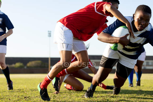 Front view of two young adult mixed race female rugby players during a match, one player running with the ball and the other reaching for the ball, with other players in the background — Stock Photo