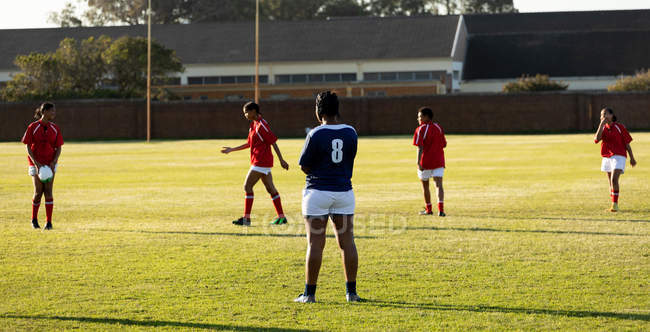 Rear view of a young adult mixed race female rugby player standing on the pitch during a rugby match, with with players from the opposing team in the background — Stock Photo