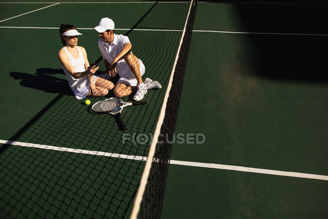 Frontansicht einer jungen kaukasischen Frau und eines Mannes, die sich an einem sonnigen Tag auf einem Tennisplatz unterhalten und ein Smartphone benutzen — Stockfoto