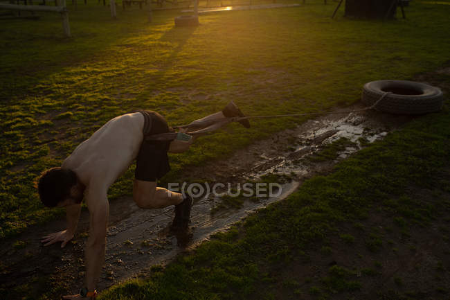 Side view of a young Caucasian man pulling a tyre on a rope around his waist through mud falling over at an outdoor gym during a bootcamp training session — Stock Photo