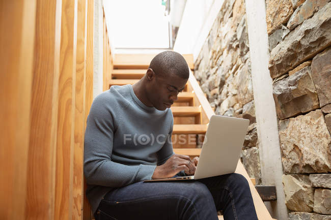Vista lateral de perto de um jovem afro-americano usando um computador portátil sentado nas escadas em casa — Fotografia de Stock