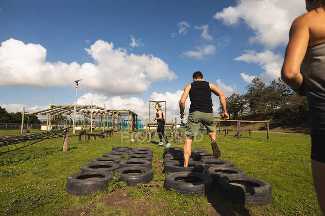 Rear view of a young Caucasian man stepping through tyres at an outdoor gym during a bootcamp training session, with one woman in the background, and one woman in the foreground waiting — Stock Photo