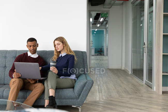 Vista frontal de un joven afroamericano y una joven mujer caucásica mirando un ordenador portátil y hablando sentado en un sofá en la sala de estar de un negocio creativo moderno - foto de stock