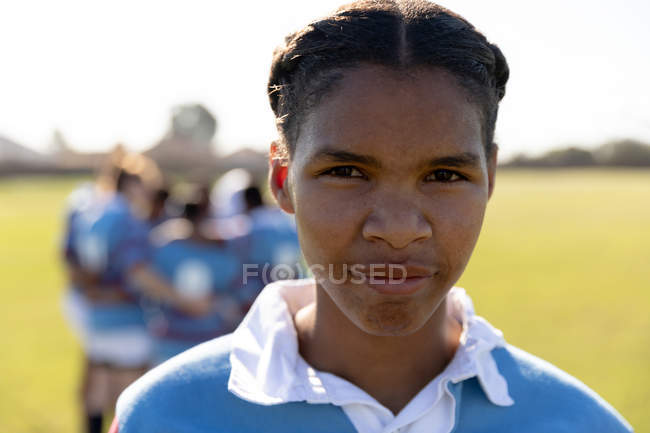 Ritratto ravvicinato di una giovane giocatrice di rugby femminile di razza mista in piedi su un campo da rugby che guarda alla telecamera, con i suoi compagni di squadra riuniti in un gruppo sullo sfondo — Foto stock