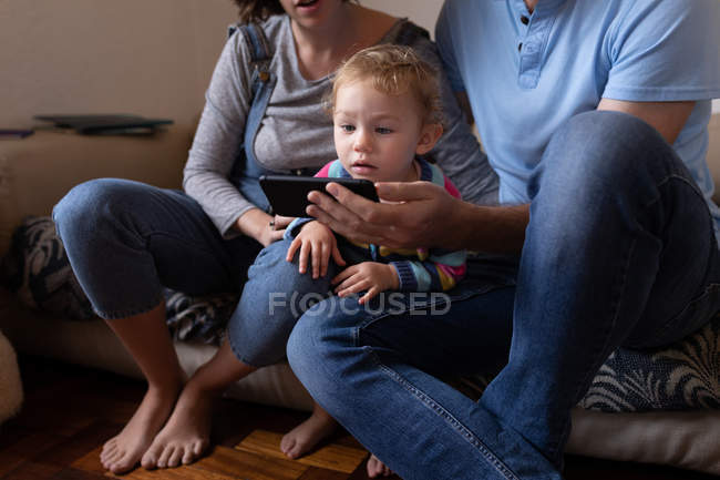 Вид спереди молодой кавказский отец и мать сидят на диване и используют смартфон со своим ребенком — стоковое фото