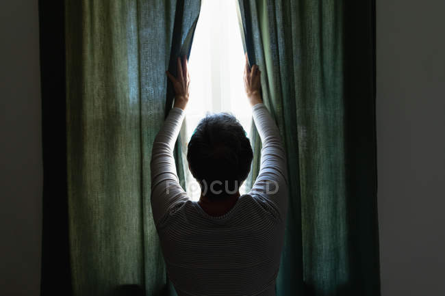 Vue arrière d'une femme blanche mature avec les cheveux courts debout et dessinant les rideaux à la maison, silhouette contre la fenêtre — Photo de stock