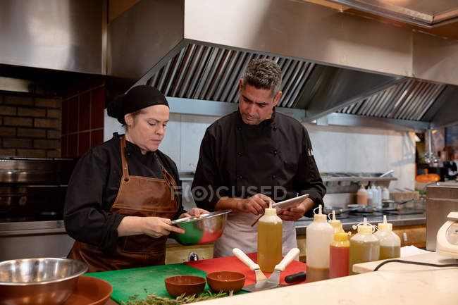 Front view close up of a middle aged Caucasian male chef holding a tablet computer and overseeing the work of a young Caucasian female chef preparing ingredients in a metal bowl in a restaurant kitchen — стокове фото