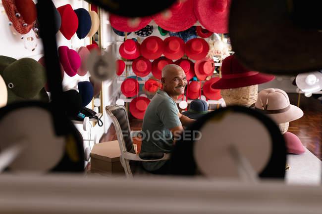 Side view of a senior mixed race man turning and smiling to camera, sitting surrounded by finished hats in the showroom at a hat factory — Stock Photo