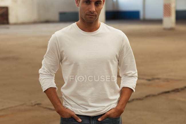 Front view close up of a young man standing with his hands in his pockets looking to camera in an empty room at an abandoned warehouse — Stock Photo