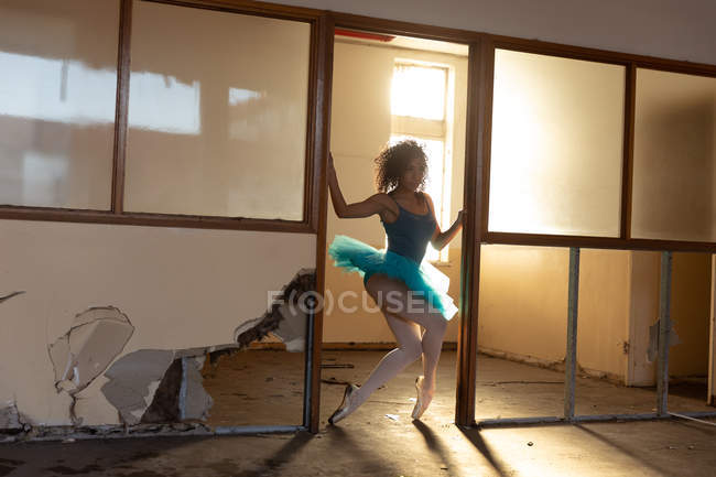 Передній вигляд молодої змішаної раси танцівниці жіночого балету в блакитному взутті, що танцює у дверному отворі в покинутій будівлі складів, підсвічування сонячним світлом — стокове фото