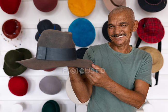 Retrato de cerca de un hombre de raza mixta mayor sonriendo y sosteniendo un sombrero terminado en la sala de exposición en una fábrica de sombreros, con varios sombreros en exhibición en el fondo - foto de stock