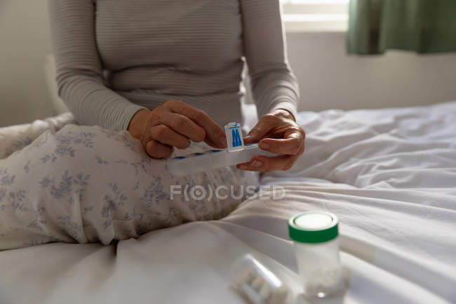 Зовнішній вигляд середині секції жінка сидить на ліжку в будинку, приймаючи ліки з щотижневої таблетки ящик, з іншими контейнерами на ліжку поруч з нею — стокове фото