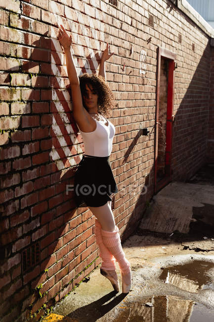 Vue de côté gros plan d'une jeune danseuse de ballet mixte debout sur ses orteils contre un mur de briques les bras levés, la tête tournée vers la caméra et les yeux fermés sur le toit d'un bâtiment urbain — Photo de stock