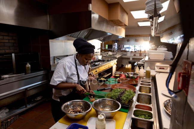Vue de côté gros plan d'une jeune chef afro-américaine hachant des ingrédients sur un comptoir dans une cuisine de restaurant occupée — Photo de stock