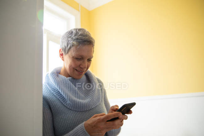 Nahaufnahme einer reifen kaukasischen Frau mit kurzen grauen Haaren, die einen Kapuzenpullover trägt, sich zu Hause mit einem Smartphone an eine Wand lehnt und lächelt, im Hintergrund das Sonnenlicht — Stockfoto