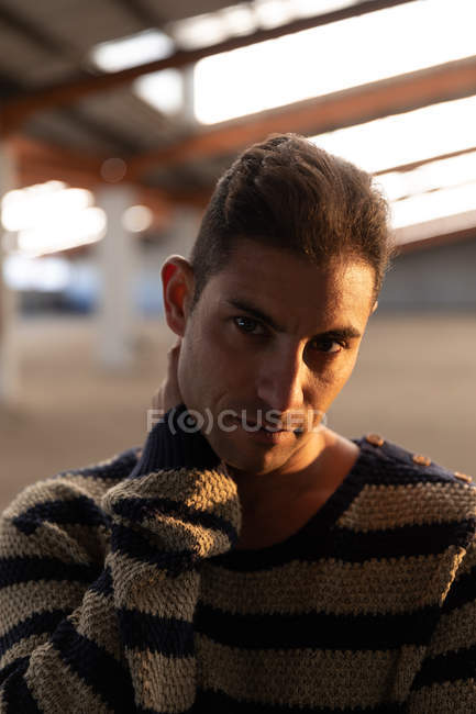Portrait close up of a young man standing with his hands around his neck and looking to camera in an empty room at an abandoned warehouse — Stock Photo