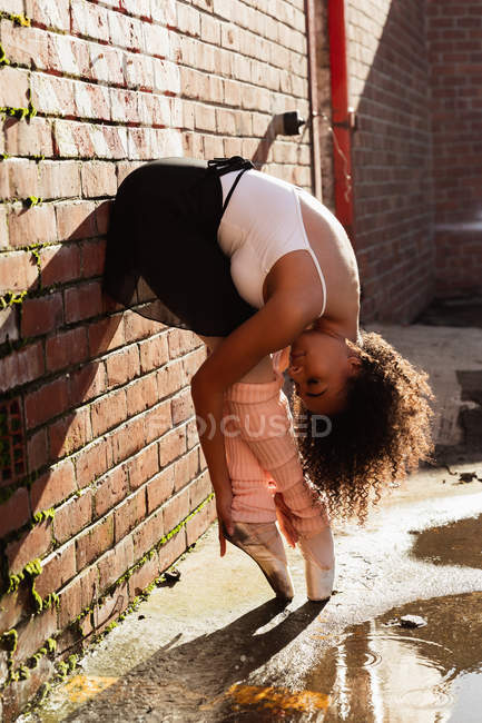 Seitenansicht einer jungen Balletttänzerin mit gemischter Rasse, die auf Zehenspitzen gegen eine Ziegelmauer steht, sich doppelt gebeugt und ihre Fersen hält, mit gedrehtem Kopf, auf dem Dach eines städtischen Gebäudes — Stockfoto