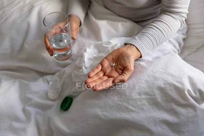 Front view mid section of woman sitting up in bed at home, holding a tablet and a glass of water, a bottle of pills on the bed beside her — Stock Photo