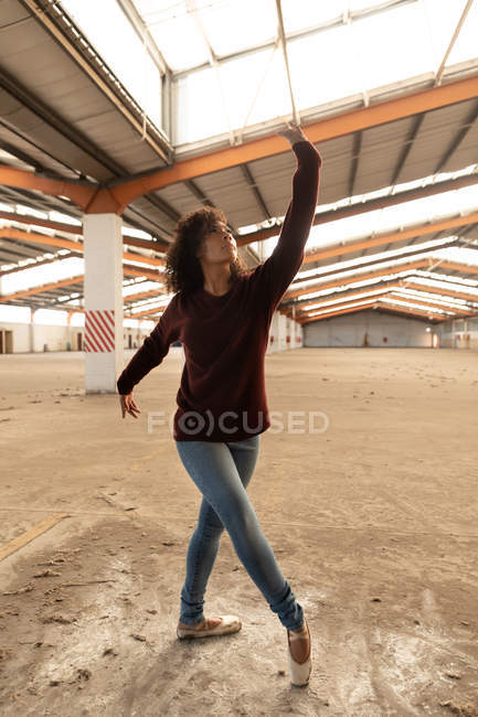 Vista frontal close up de uma jovem mestiça dançarina de balé vestindo jeans e sapatos pontiagudos dançando com os braços estendidos em um armazém abandonado — Fotografia de Stock