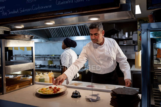 Вид спереди на белого повара средних лет, готового подавать блюда на кухне ресторана, в то время как мимо него проходит женщина из персонала кухни — стоковое фото