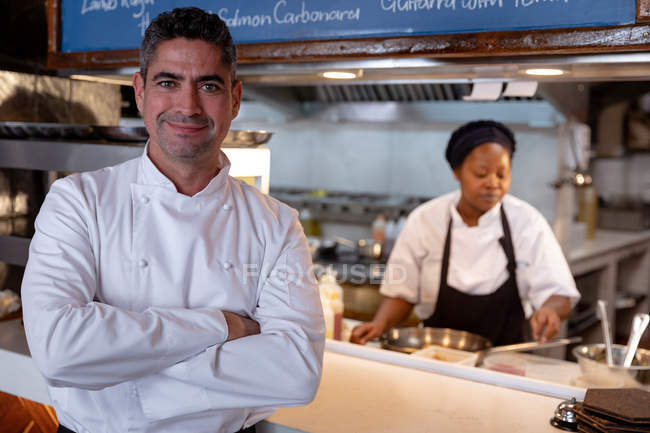 Vista frontal de cerca de un chef caucásico de mediana edad de pie con los brazos cruzados sonriendo a la cámara, mientras que una joven chef afroamericana está detrás de él trabajando en una cocina de restaurante - foto de stock