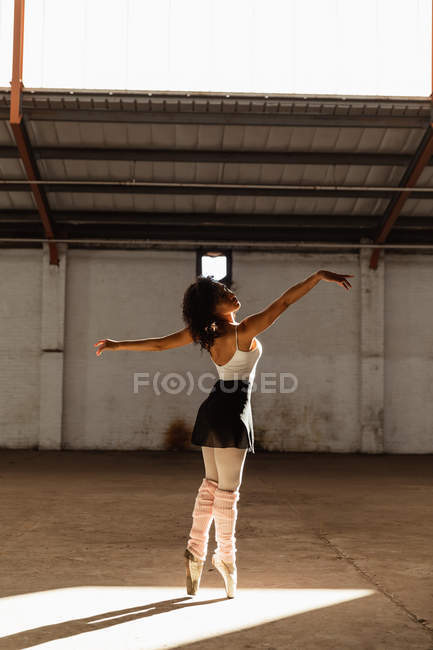 Vista laterale di una giovane ballerina di danza mista che indossa scarpe da punta in piedi sulle dita dei piedi sotto il sole con le braccia tese mentre balla in una stanza vuota in un magazzino abbandonato — Foto stock