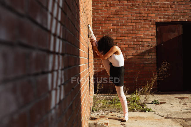 Veduta laterale di una giovane ballerina di danza mista in piedi su una gamba con le dita dei piedi che tiene l'altra gamba contro un muro di mattoni con entrambe le braccia, sul tetto di un edificio urbano — Foto stock
