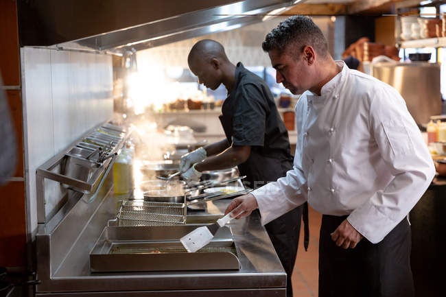 Side view close up of a middle aged Caucasian male chef and young African American male member of the kitchen staff standing over fryers in a restaurant kitchen — Stock Photo