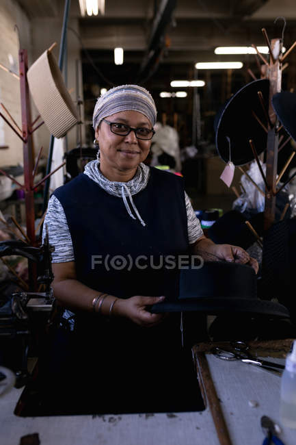 Портрет крупным планом смешанной расы средних лет женщины, стоящей за столом держа шляпу в мастерской на шляпном заводе, в окружении оборудования и материалов — стоковое фото