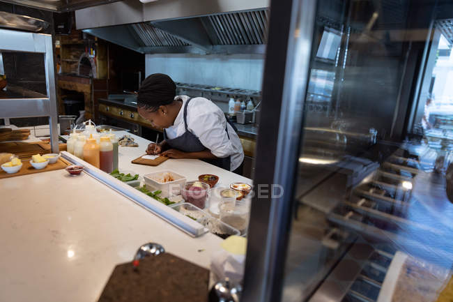 Vista lateral de una joven chef afroamericana escribiendo una nota en el mostrador en la cocina de un restaurante - foto de stock