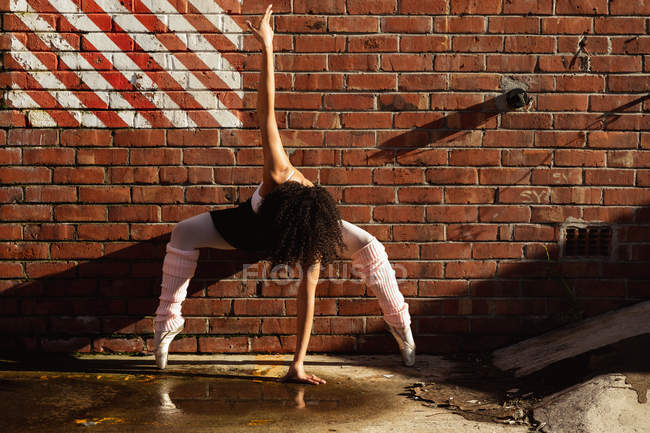 Вид спереду молода змішана гонка танцівниця балету, що стоїть на пальцях на цегляній стіні з піднятою однією рукою і однією рукою торкається землі, на даху міської будівлі — стокове фото