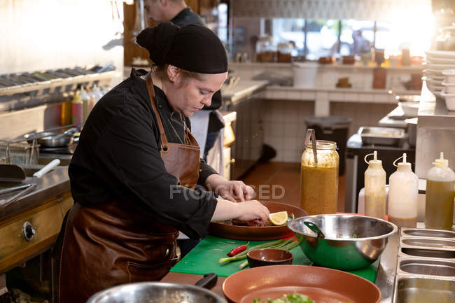 Vue de côté gros plan d'une jeune cuisinière caucasienne travaillant à un comptoir préparant un plat dans une cuisine de restaurant — Photo de stock