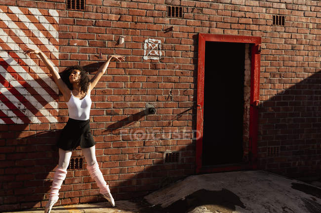Вид спереду молода змішана раса танцівниця балету, що стоїть на пальцях на цегляній стіні з піднятими руками і дивитися на дах міської будівлі — стокове фото