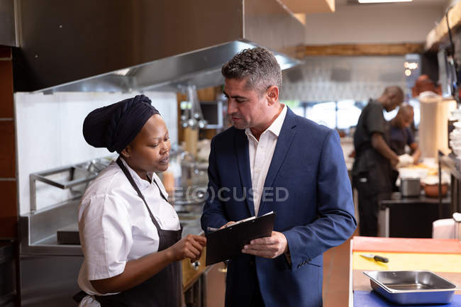 Vista frontal de cerca de un gerente de restaurante caucásico de mediana edad sosteniendo un portapapeles y hablando con una joven chef afroamericana en una cocina de restaurante, con el personal de cocina trabajando en el fondo - foto de stock