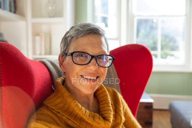 Ritratto ravvicinato di una donna caucasica matura dai capelli corti e grigi con occhiali e maglione a collo alto, seduta in una poltrona rossa nel suo salotto guardando la macchina fotografica e sorridendo, una finestra illuminata dal sole sullo sfondo — Foto stock