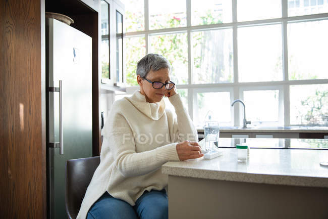 Vue latérale d'une femme caucasienne mature aux cheveux gris court portant des lunettes assise dans sa cuisine lisant sur ses médicaments, avec des bouteilles de pilules, une boîte à pilules hebdomadaire et un verre d'eau sur le comptoir à côté d'elle — Photo de stock