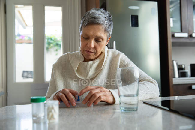 Вид спереду крупним планом зрілої кавказька жінка з коротким сивим волоссям сидить у своїй кухні, дивлячись на її ліки, з таблетках, щотижневі таблетки і склянку води на прилавку поруч з нею — стокове фото