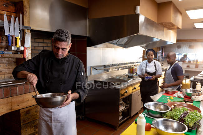 Вид спереду крупним планом середнього віку Кавказького чоловічого шеф-кухаря змішування інгредієнтів в металеву миску в зайнята кухні ресторану, з іншими працівниками кухні, що працюють разом у фоновому режимі — стокове фото